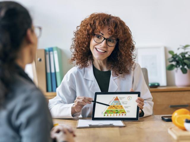 Nutricionista explicando la pirámide alimenticia a una paciente con una tableta digital en consulta médica.